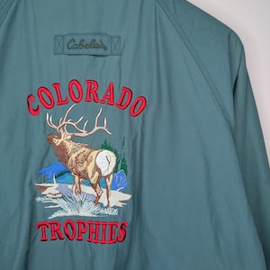 Puede incluir: Una chaqueta de color verde azulado con cuello gris y un diseño bordado en la espalda. El bordado incluye el texto Cabela's, la palabra COLORADO en rojo, una ilustración de un alce en un paisaje de montaña y la palabra TROPHIES en letras rojas.