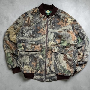 Könnte beinhalten: Eine Camouflage-Bomberjacke mit braunem Reißverschluss, Bündchen und Kragen. Die Jacke hat zwei große Fronttaschen und einen kleinen Logoaufnäher auf der Brust. Das Muster umfasst Brauntöne, Grün und Beige.