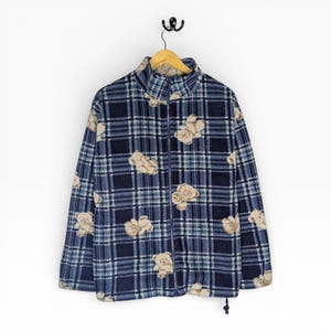 May include: A blue fleece jacket with a plaid pattern and teddy bear graphics. The jacket has a full zipper, a high collar, and long sleeves. It is hanging on a wooden hanger against a white background.