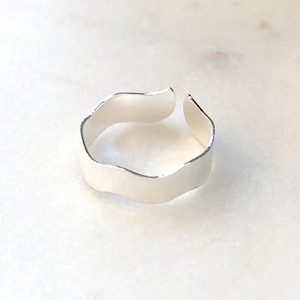 Könnte beinhalten: Ein verstellbarer Ring in Silber mit einem wellenförmigen, organischen Design. Der Ring ist oben offen, um die Größe anzupassen. Das Metall hat eine glatte, reflektierende Oberfläche, und der Ring ist vor einem weißen Marmorhintergrund platziert.