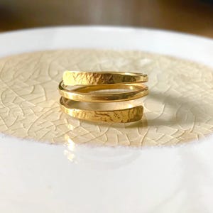 Könnte beinhalten: Ein goldfarbener Ring mit dreifachem Wickeldesign und einer texturierten, gehämmerten Oberfläche. Das Band besteht aus drei überlappenden Windungen, die ein geschichtetes Aussehen erzeugen und ein metallisches Finish mit sichtbaren Vertiefungen.