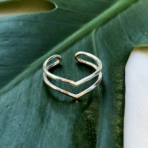 Könnte beinhalten: Ein silberfarbener, verstellbarer Ring mit doppeltem Chevron-Design. Der Ring hat zwei V-förmige Bänder, die nebeneinander liegen und einen geschichteten Effekt erzeugen. Der Ring wird auf einem dunkelgrünen Blatt gezeigt.