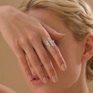 Könnte beinhalten: Ein silberner Ring mit zwei parallelen vertikalen Stäben. Der Ring wird an einem Finger getragen, wobei die Hand das Gesicht teilweise verdeckt. Die Nägel sind in einem neutralen Farbton lackiert. Der Hintergrund ist ein warmes Beige.