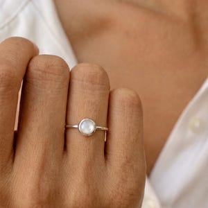 Könnte beinhalten: Ein Silberring mit einem einzelnen, runden, in einer Zargenfassung gefassten Mondstein-Cabochon. Der Edelstein zeigt einen sanften, irisierenden weißen Schimmer, während die Ringschiene aus poliertem, glattem Silbermetall gefertigt ist.