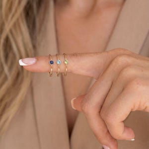 Könnte beinhalten: Drei dünne Goldringe, jeder mit einem einzelnen kleinen runden Edelstein in einer Fassung. Die Steine sind dunkelblau, hellblau und blassgelb und in einer Reihe an einem Finger angeordnet. Die Ringbänder sind schlicht, glatt und goldfarben.