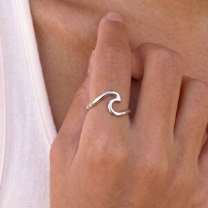Könnte beinhalten: Ein Ring aus poliertem Silber mit einem durchgehenden, fließenden Wellendesign, das sich um den Finger legt. Das Band ist glatt und metallisch und zeigt eine minimalistische, offene Form, die den Kamm einer Meereswelle nachahmt.
