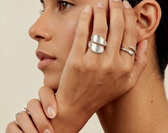 May include: A silver-toned bypass ring with a smooth, polished finish on the middle finger, paired with a slender, textured silver-toned ring on the ring finger. Additional thin silver-toned rings are visible on the fingers of the other hand.