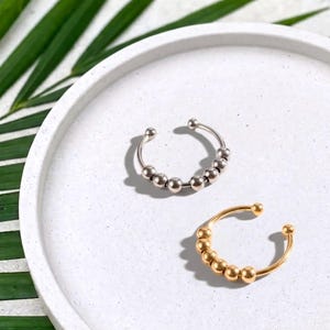 Könnte beinhalten: Zwei offene Ringe, einer in Silberoptik und einer in Goldoptik, die jeweils mit einer Reihe kleiner, glatter, beweglicher Metallperlen entlang der Mitte des Rings versehen sind. Die Ringe sind kreisförmig mit einer Öffnung an der Oberseite.