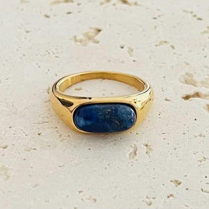 Könnte beinhalten: Ein goldfarbener Ring mit einer glatten, polierten Ringschiene und einem mittig eingesetzten, horizontal ausgerichteten ovalen blauen Stein. Der Stein zeigt tiefe marineblaue Farbtöne mit subtilen, natürlichen Farb- und Strukturvariationen.