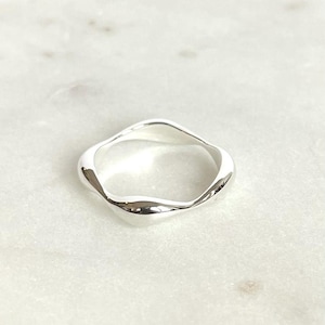 Könnte beinhalten: Ein silberner Ring mit einem einzigartigen, wellenförmigen Design. Der Ring hat eine glatte, polierte Oberfläche und eine moderne, minimalistische Ästhetik. Die Form des Rings ist ein unregelmäßiges Quadrat mit weichen, gebogenen Kanten.