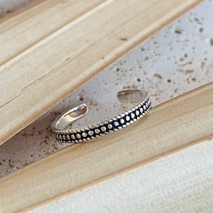 Könnte beinhalten: Ein silberner Zehenring mit gepunktetem Design. Der offene Ring besteht aus glänzendem Metall und weist eine Reihe kleiner, gleichmäßig verteilter Punkte um das Band auf. Der Ring wird auf einer hellen Oberfläche mit Holzmaserung präsentiert.