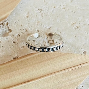 Könnte beinhalten: Ein silberner Zehenring mit einer Reihe kleiner, gleichmäßig verteilter, runder Perlen. Der Ring ist oben offen, um ihn anzupassen. Der Ring wird auf einer hellen, strukturierten Oberfläche mit einem getrockneten Blatt gezeigt.