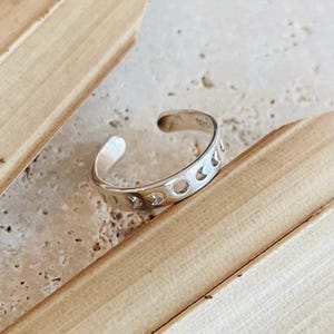 Può includere: Un anello da dito del piede color argento con un design a mezzaluna. L'anello ha una forma curva ed è aperto nella parte superiore. L'anello è su una superficie chiara con una trama naturale e organica.