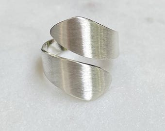 May include: A silver-toned bypass ring featuring a wide, open-ended band with a brushed metal finish. The design consists of two overlapping, tapered ends that curve around the finger, creating a continuous, fluid shape with a matte texture.