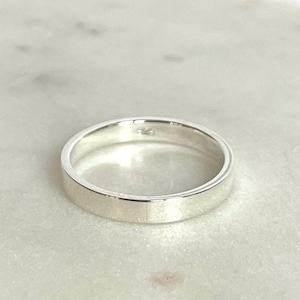 Könnte beinhalten: Nahaufnahme eines silbernen Rings mit flachem Band. Der Ring besteht aus glänzendem, reflektierendem Metall und liegt auf einer hellen Oberfläche. Das Design des Rings ist schlicht und elegant, für den Alltag geeignet.