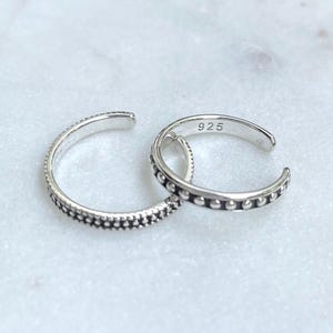 Könnte beinhalten: Zwei silberne Zehenringe. Ein Ring hat ein gepunktetes Design, der andere ein Perlenmuster. Die Ringe sind aus glänzendem Metall gefertigt und werden auf einer weißen Marmoroberfläche präsentiert. Die Zahl "925" ist auf einen der Ringe gestempelt.