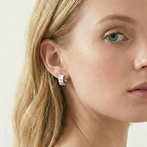 Könnte beinhalten: Silberne Creolen mit einem strukturierten, geometrischen Design. Die Ohrringe sind klein und schmiegen sich an das Ohrläppchen an. Das Bild zeigt eine Nahaufnahme der Ohrringe an einem Ohr.