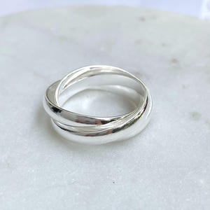 Könnte beinhalten: Ein polierter Silberring mit einem ineinandergreifenden Design aus drei Bändern. Die drei glatten, abgerundeten Ringe sind miteinander verwoben und bilden ein zusammenhängendes Schmuckstück mit einer reflektierenden, metallischen Oberfläche.