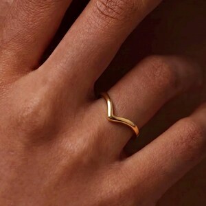 Könnte beinhalten: Ein polierter Goldring mit einem glatten, V-förmigen Chevron-Design. Das Band hat ein abgerundetes Profil und eine durchgehende, massive metallische Oberfläche, die einer sanften, winkligen Kontur über den Finger folgt.