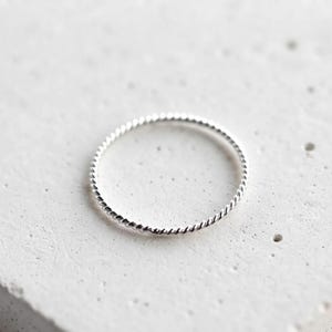 Könnte beinhalten: Ein dünner, kreisförmiger Ring aus poliertem, silberfarbenem Metall. Das Band weist eine durchgehende, feine gedrehte Seilstruktur auf, die sich um den gesamten Umfang des Rings legt und ein gleichmäßiges, sich wiederholendes Spiralmuster bildet.