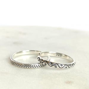 Könnte beinhalten: Zwei Ringe aus 925er Sterlingsilber. Ein Ring weist ein geflochtenes Texturmuster um das Band auf. Der zweite Ring zeigt ein sich wiederholendes Wellendesign mit kleinen kreisförmigen Vertiefungen in der Mitte und ist innen mit 925 markiert.