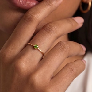 Könnte beinhalten: Ein zarter Goldring mit einem kleinen, runden, leuchtend grünen Edelstein. Der Ring hat ein schmales Band und ein schlichtes, elegantes Design. Der Edelstein ist in einem minimalistischen Stil gefasst, der seine lebendige Farbe hervorhebt. Der Ring ist ein Schmuckstück.