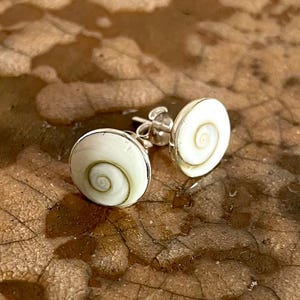 Könnte beinhalten: Ein Paar Ohrstecker aus Silber. Jeder Ohrring hat ein weißes, spiralförmiges Muscheldesign mit silbernem Rand. Die Ohrringe sind vor einem braunen, strukturierten Hintergrund platziert.