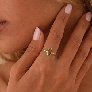 Könnte beinhalten: Ein goldener Ring mit einem braunen, marquiseförmigen Edelstein. Der Ring wird auf einer Hand mit rosa Nagellack gezeigt. Das Ringband ist schmal, und der Edelstein ist in einem schlichten Design gefasst. Schmuck.