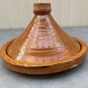 May include: A brown ceramic tagine lid with a wide, flat base and a conical top. The lid has a glazed finish and a slightly raised rim.