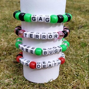 May include: Four colorful beaded bracelets stacked on a white cylinder. The bracelets feature black, green, pink, and red beads, with white letter beads spelling out words like "GAG", "TERROR", "FREE BUGS", and "BIG DEATH".