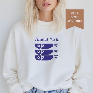 May include: White crewneck sweatshirt with a blue graphic of three stacked fish and the text "Tinned Fish".