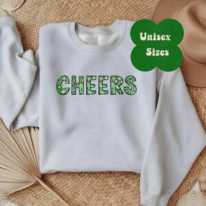 May include: A light gray sweatshirt with the word "CHEERS" printed in green shamrock-patterned letters. The sweatshirt is available in unisex sizes.