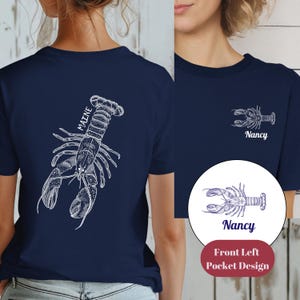 May include: A navy blue t-shirt with a white lobster design on the back and a smaller lobster design on the front left pocket. The text "MAINE" is written vertically on the back of the shirt. The text "Nancy" is written below the lobster design on the front pocket.