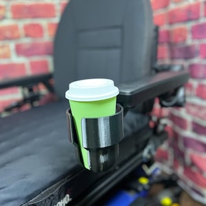 May include: A black 3D-printed cup holder attached to a wheelchair armrest, holding a green coffee cup with a white lid. The wheelchair has a black seat and backrest, with a brick wall background.