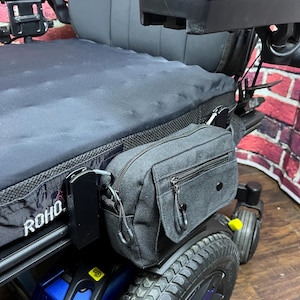 May include: Black wheelchair with a black seat cushion and armrests. A black bag with a zipper and a small pocket is attached to the side. The wheelchair has a blue and black wheel with a black tire. The word "ROHO" is visible on the seat.