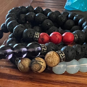 May include: A set of five bracelets made of black lava rock beads, amethyst beads, red beads, light brown beads, and white beads. Each bracelet has silver metal accents.