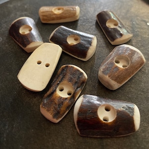May include: A collection of eight antler buttons in various shades of brown and beige. Some buttons have two holes, while others have four holes.