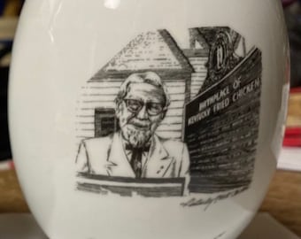 May include: White ceramic mug with a black and white illustration of Colonel Harland Sanders, the founder of Kentucky Fried Chicken. The illustration shows him standing in front of a building with the text "Birthplace of Kentucky Fried Chicken".
