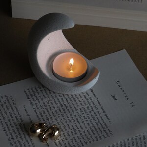 Może przedstawiać: Szary, betonowy świecznik w kształcie półksiężyca z zapaloną świeczką typu tealight. Złote kolczyki koła spoczywają na otwartej książce. Scena przywołuje poczucie spokoju i relaksu.