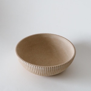 Può includere: Una ciotola in ceramica beige con un esterno strutturato e a coste. La ciotola ha una forma arrotondata e un aspetto maculato. Il bordo della ciotola ha un bordo decorativo smerlato. La ciotola è vuota e poggia su una superficie bianca.
