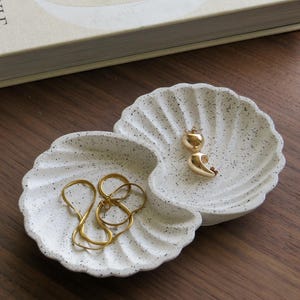 May include: A double shell-shaped trinket dish made of off-white material with black speckles. The dish features two scalloped compartments holding gold-toned jewellery, including a pair of small hoop earrings and a thin gold chain necklace.