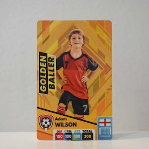 May include: A yellow, orange, and black card with a photo of a young boy wearing a red and black soccer jersey. The card reads "Golden Baller" and "Adam Wilson". The card also includes a soccer ball graphic and a scorecard with the categories "Def", "Ctrl", "Att", and "Total".