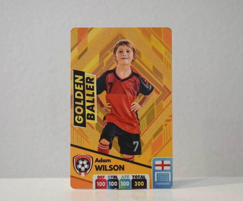 May include: A yellow, orange, and black card with a photo of a young boy wearing a red and black soccer jersey. The card reads "Golden Baller" and "Adam Wilson". The card also includes a soccer ball graphic and a scorecard with the categories "Def", "Ctrl", "Att", and "Total".