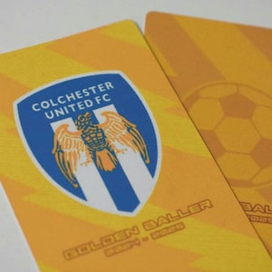 May include: Two yellow and orange cards with the text "Colchester United FC" and a blue and white crest with a bird. The text "Golden Baller" is on the bottom of the card.