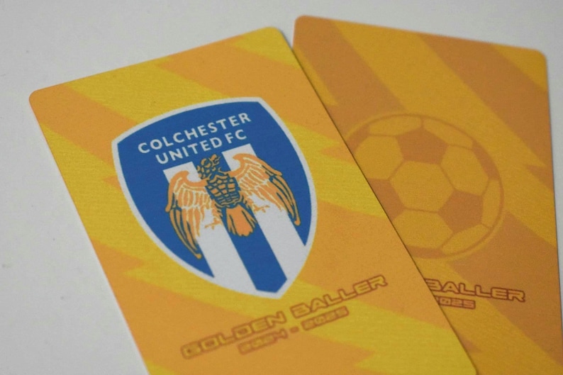 May include: Two yellow and orange cards with the text "Colchester United FC" and a blue and white crest with a bird. The text "Golden Baller" is on the bottom of the card.