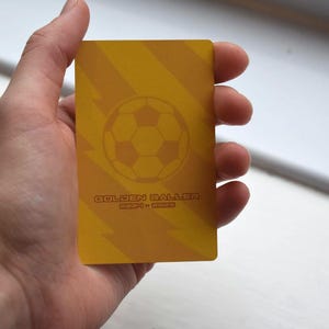 May include: A yellow card with a soccer ball graphic and the text "Golden Baller" in red. The card has a striped pattern.