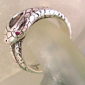 May include: A silver snake ring with a ruby red eye. The snake is coiled and the tail wraps around the finger.