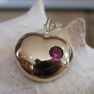 Może przedstawiać: Złoty wisiorek w kształcie serca z pojedynczym różowym kamieniem szlachetnym osadzonym w środku.