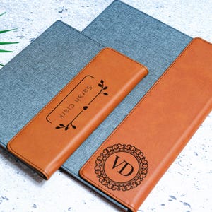 May include: Two brown leather journals with personalized engravings. The larger journal has the initials "VD" in a decorative circle. The smaller journal has the name "Sarah Clark" with a floral design.