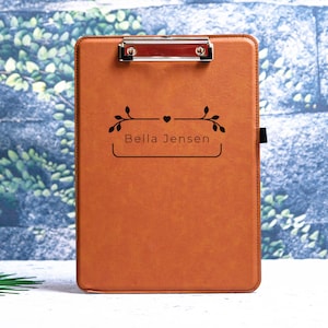 May include: A brown leather clipboard with a silver clip and a personalized inscription that reads "Bella Jensen" with a small heart and floral design.